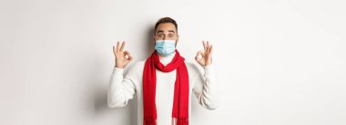 Covid-19, tecrit ve karantina konsepti. Yüz maskeli etkileyici bir adam, tamam işaretleri gösteriyor ve katılıyor, harika bir ürün öneriyor, beyaz arka plan..