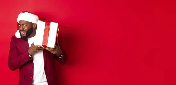 Christmas. Curious Black man in santa hat shaking new year gift, wonder what inside box, standing against red background.