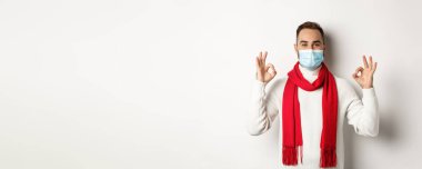 Covid-19, tecrit ve karantina konsepti. Yüz maskesi takmış tatmin olmuş yakışıklı bir adam işaret veriyor, onaylıyor ya da kabul ediyor..