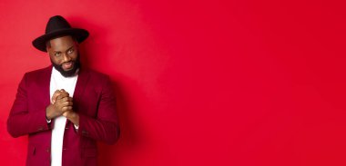 Kurnaz siyah adamın bir fikri var, gülümsemesi sinsi ve ellerini ovuşturuyor, kırmızı arka plana karşı duruyor ve entrika çeviriyor..