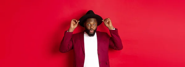 Fashion and party concept. Handsome Black man wearing hat going on holiday celebration, looking excited, standing over red background.