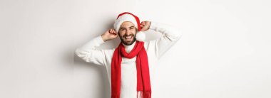 Kış tatili ve yeni yıl partisi konsepti. Noel 'i kutlayan mutlu adam, Noel Baba şapkası ve kırmızı eşarp takıyor, yüksek sesle gülüyor, beyaz arka plan.