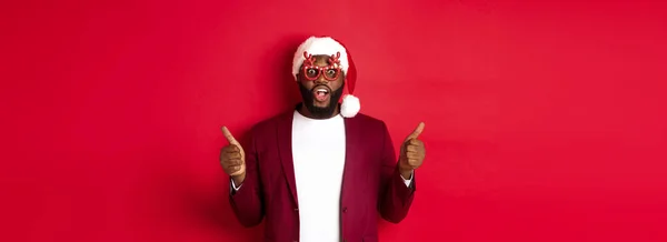 Funny Black man celebrating New Year, wearing party glasses and santa hat, showing thumb up, approve and like, standing over red background.