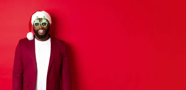 Funny Black man in santa hat and party glasses celebrating Christmas, smiling happy and wishing merry xmas, standing over red background.