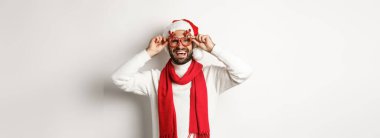 Noel, yeni yıl ve kutlama konsepti. Mutlu adam gülüyor, Noel Baba şapkası ve parti gözlüğü takıyor, beyaz arka planda duruyor..