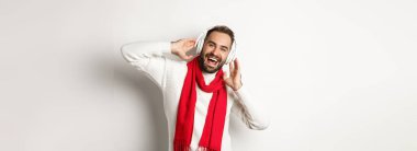 Kış tatili ve teknoloji konsepti. Mutlu adam gülümsüyor, kulaklıkla müzik dinliyor, süveter takıyor, beyaz arka plan.