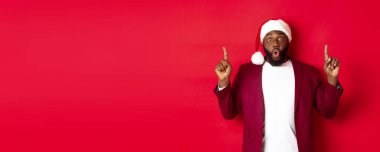 Noel, parti ve tatil konsepti. Etkilenmiş siyahi adam teklif gösteriyor, parmaklarını logoya doğrultuyor, vay canına diyor, kırmızı arka planda duruyor..