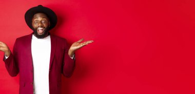 Noel alışverişi ve insanların konsepti. Yakışıklı Afrikalı Amerikalı adam gülümsüyor, ellerini yana açıyor, fotokopi alanında tanıtım teklifleri gösteriyor, kırmızı arka plan..