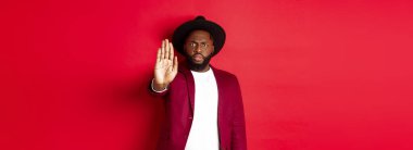 Ciddi ve endişeli siyahi adam seni durdurmak için elini uzatıyor, yasaklıyor ve karşı çıkıyor, somurtuyor ve surat asıyor, kırmızı arka planda dikiliyor..