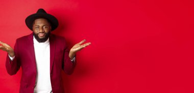 Cahil ve umursamaz siyah adam kayıtsız bakışlarla omuz silkiyor, umursamıyor, kırmızı arka planda duruyor..