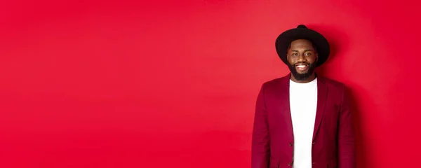 Fashion and party concept. Stylish african american man in blazer and hat, smiling confident at camera, celebrating new year, standing over red background.