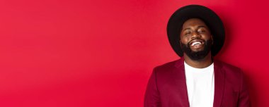 Siyah şapkalı, ceketli, kırmızı arka planlı, mutlu ve yakışıklı siyah adam sana gülümsüyor..