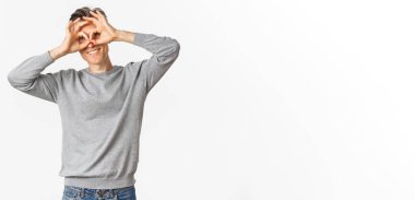 Orta yaşlı komik bir adamın portresi, yüz ifadeleri yapıyor, el gözlüklerini gözlerinin etrafına gösteriyor, sanki dürbünle bakıyor gibi, beyaz arka planda duruyor..