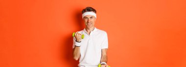 Yakışıklı orta yaşlı erkek sporcunun görüntüsü, dambıllarla egzersiz, gülümseme, turuncu arka planda antrenman..