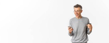 Gri süveter ve gözlüklü, kahve içen ve akıllı telefon ekranında mutlu görünen, internette bir şeyler okuyan, beyaz arka planda duran orta yaşlı yakışıklı bir adam..