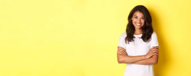 Beyaz tişörtlü, göğsünde çapraz kolları olan ve sarı arka planda duran mutlu, Afro-Amerikan bir kız resmi..