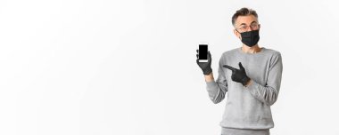 Covid-19 kavramı, sosyal mesafeler ve yaşam tarzı. Gözlüklü, tıbbi maskeli ve eldivenli çekici orta yaşlı bir adamın görüntüsü bir şeyi açıklıyor ve cep telefonu ekranını işaret ediyor..