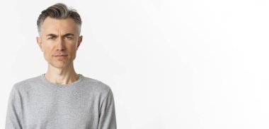 Şüpheci orta yaşlı adamın yakın çekimi, kaşlarını çatıyor ve kameraya şüpheli bakıyor, gri süveter giyen beyaz arka planda duruyor..