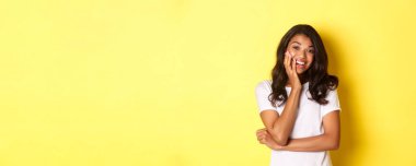 Afro-Amerikalı çekici bir kızın portresi, şaşırmış ve gülümsüyor, sarı arka planda sıradan beyaz tişörtle dikiliyor..