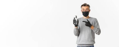 Koronavirüs, yaşam tarzı ve karantina kavramı. Medikal maskeli orta yaşlı bir adam resmi ve eldivenler el dezenfektanı kullanmanızı tavsiye ediyor, antiseptik şişeyi işaret ediyor, beyaz arka planda duruyor..