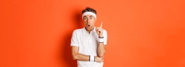 Spor, spor ve yaşam tarzı kavramı. Yakışıklı orta yaşlı erkek sporcunun portresi, bir fikri var, parmağını kaldırıyor ve öneriyor, turuncu arka planda duruyor..