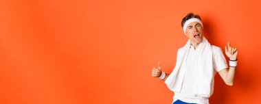 Mutlu ve kaygısız orta yaşlı fitness elemanı, antrenmandan sonra dans eden, havluyu boynuna dayayan, turuncu arka planda poz veren.