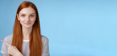 Çekici, mütevazı, güzel, kızıl saçlı olgun, dostane, gülümseyen, sol işaret parmağıyla işaret eden, yol gösteren, banyoya bakan, mavi arka planda mutlu bir sırıtış..