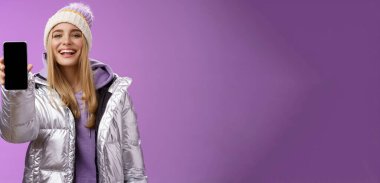 Şımarık, neşeli, sarışın bir kadın elinde cep telefonu görüntüsü, gururla tatmin edici gülümseme kamerası, ayakta duran mor arkaplan gösteren akıllı telefonları kullanarak uygulamayı düzenlemeyi öneriyor..