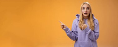 Kapüşonlu, sarışın, şaşkın bakışlı, şaşırmış, şaşırmış bakışlı, ağzı açık, solu işaret eden, dedikoducu, turuncu arka planda duran sarışın kadın şaşırmış..