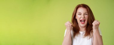 Evet, başardık. Neşeli, şanslı, kızıl saçlı, orta yaşlı, kadın kazanan yumrukları çakıyor kutlama başarısı jesti bağırıyor, sevinç çığlıkları atıyor harika haberleri kutluyoruz yeşil arka plan mutlu.