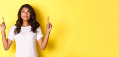 Çekici Afro-Amerikan kadın modelin portresi, beyaz tişört giyiyor, meraklı bir gülümsemeyle parmaklarını kaldırıp bakıyor, reklamını yapıyor, sarı arka planda duruyor..