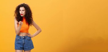 Kadın seksi erkeği baştan çıkarıyor. Şehvetli ve seksi, kıvırcık saçlı, modern kesimli bluzlu ve şortlu, çekici, çekici, çekici, turuncu arka planda flörtleşen kadın..