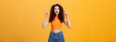 Kendine güvenen beyaz bir kadının portresi. Kırmızı rujlu kıvırcık saç stili ve turuncu üst kısmı kaldırılmış ellerle yukarı bakıyor ve kameraya neşeyle gülümsüyor..