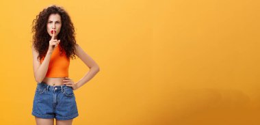 Kardeş hoşnutsuz sözler söylüyor ciddi ve katı sözler söylüyor turuncu arka planda şık kıyafetler içinde kameraya şşşş diyor ağzında işaret parmağıyla itaat kuralları talep ediyor.