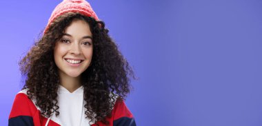 Yumuşak, kıvırcık saçlı, 20 'li yaşlarda, kalın bereli ve havalı tişörtlü harika güneşli ve serin günlerin tadını çıkartan, mavi arka planda poz veren güzel bir kadın. Boşluğu kopyala