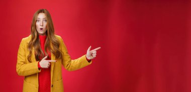 Dili tutulmuş, kafası karışmış, sarı sonbahar ceketli, ağzı açık kırmızı saçlı, şaşırmış bir şekilde kırmızı duvarın üzerinde şaşkın ve habersiz bir şekilde kameraya bakan bir kız.