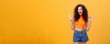 Cana yakın ve arkadaş canlısı beyaz bir kız. Kıvırcık saçlı, kısa şortlu. Onaylı jest yapıyor. Turuncu duvarın üstünde kameraya gülümseyen muhteşem partileri sevdiğini söylüyor..