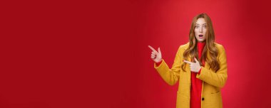 Dili tutulmuş ve etkilenmiş, sarı ceketli, çilli, çekici, kızıl saçlı kadın. Çene, kamera karşısında şaşkın şaşkın, sorgulanan gözler, kırmızı arka plan üzerinde sol işaret eden..