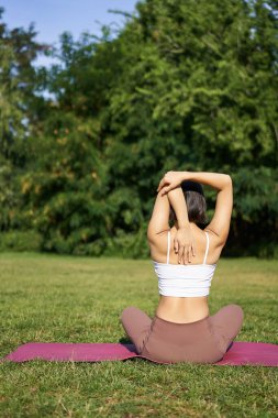 Sporcunun antrenmanın ortasında arka tarafa bakması, amrs arkasını germesi, parkta çimenlikte kauçuk yoga minderinde oturması..