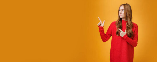 Studio shot of suspicious serious-looking unsure redhead girl frowning looking and pointing at upper left corner with doubtful strict gaze posing uncertain over orange background. Copy space