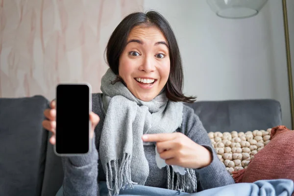 Excited young woman pointing finger at smartphone, showing online doctor, medical application or GP contact on mobile phone, staying at home sick, catching cold.