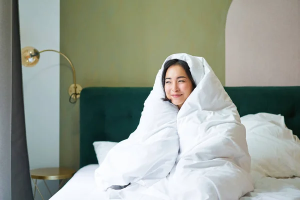 Cozy mornings. Happy asian girl feels warm in her bed, covers herself with cozy duvet in her bedroom, a comfortable stay in hotel room.