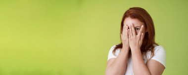 Dikizlemiyorum. Karizmatik orta yaşlı kızıl saçlı kadın avuçlarının içine bak parmaklarının arasından bak bir göz kamerasının ilgisini çek sürpriz bir hediye bekle yeşil arka plan.