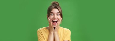 Sarı tişörtlü mutlu ve şaşırmış genç bir kızın portresi şaşkınlık ve sevinçten yanaklarına bastırıyor yeşil arka plandaki hayret verici habere geniş bir gülümsemeyle tepki veriyor.