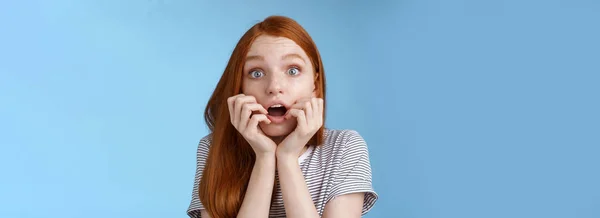 Shocked speechless gasping young redhead girl staring impressed stunned watching important moment tv series biting fingers open mouth shook standing excited blue background anticipating.