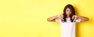 Şüpheci, eğlendirilmemiş, Afro-Amerikan kızı tasviri, baş parmağını kaldırıyor ve yüzünü buruşturuyor, kötü bir şeyi yargılıyor, sarı arka planda hayal kırıklığına uğruyor..