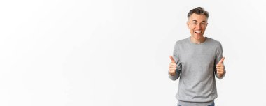 Şaşırmış ve mutlu orta yaşlı bir adamın resmi, başparmaklarını yukarı kaldırıyor ve gülümsüyor, beyaz arka planda duruyor..