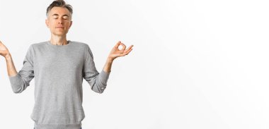 Sakin ve rahat orta yaşlı bir adamın portresi, rahatça nefes alıyor, gözleri kapalı meditasyon yapıyor ve elleri yana doğru açık, beyaz arka planda yoga yapıyor..