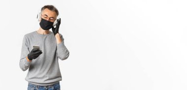 Covid-19 kavramı, sosyal mesafeler ve yaşam tarzı. Medikal maskeli, eldiven ve gözlüklü, kulaklıkla müzik dinleyen ve elinde akıllı telefon tutan orta yaşlı bir adam resmi.