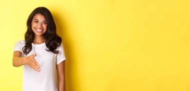 Çekici Afro-Amerikan kız portresi gülümseyen, el sıkışmak için uzanan, seni selamlayan, sarı arka planda duran.